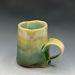 May include: Handmade ceramic mug with a textured, vertical ribbed design. The mug features a green and yellow glaze, with a rounded handle. The mug is suitable for coffee, tea, or other beverages.