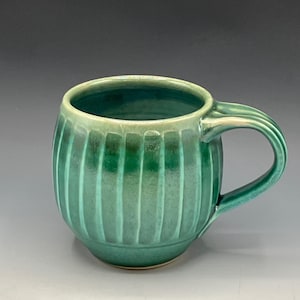 May include: A handmade ceramic mug with a ribbed design and a glossy, teal-green glaze. The mug has a rounded body, a curved handle, and a light green rim. This unique mug is perfect for enjoying coffee, tea, or other beverages.