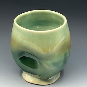 May include: A handmade ceramic cup with a unique design. The cup features a rounded shape with a hole on the side and a small base. The color gradient transitions from light green to a darker teal, with a touch of yellow at the base.