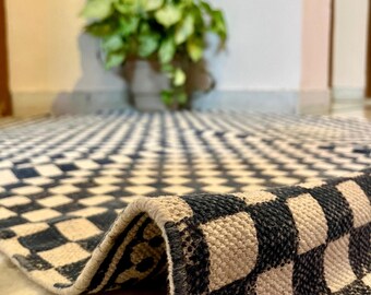 May include: A woven area rug featuring a repeating black and off-white checkerboard pattern. The rug is partially folded, revealing the textured weave and finished edges. A small logo with the text Samyuktha Handicrafts is visible.