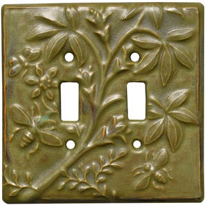 May include: A square olive green ceramic double toggle light switch plate featuring a raised relief design of botanical branches, leaves, a small flower, and two bees. The plate has four screw holes and two rectangular openings for switches.