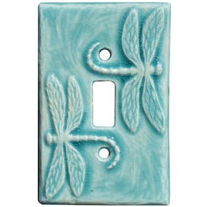 Puede incluir: Cubierta de interruptor de luz de cerámica turquesa con acabado craquelado. Dos diseños de libélulas blancas en relieve. Tiene una abertura rectangular para el interruptor y dos orificios para tornillos.