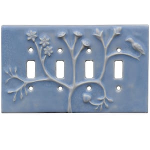 May include: A pale blue ceramic four-gang light switch plate featuring a raised design of a tree and a bird. The tree branches and flowers are white. It has four rectangular switch openings and four small screw holes.