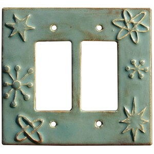 May include: A turquoise ceramic double light switch cover with raised star and floral designs. The square plate has a distressed finish and four screw holes. The decorative plate adds a unique touch to any room.