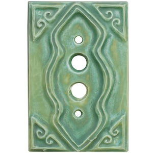 Może przedstawiać: Prostokątna, zielona ceramiczna płytka do włącznika światła z centralnym, wydłużonym wzorem z trzema okrągłymi otworami. Płytka ma teksturowaną powierzchnię i dekoracyjne zawijasy w rogach.