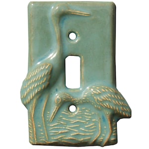 Puede incluir: Una placa de interruptor de luz rectangular de cerámica azul claro con un diseño en relieve de dos aves de cuello largo. Un ave está de pie a la izquierda mientras la otra se inclina hacia abajo a la derecha. La placa tiene dos orificios para tornillos y una abertura central.