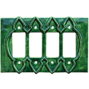 以下が含まれることがあります： 光沢のあるエメラルドグリーンのセラミック製スイッチプレート。4つの長方形の開口部があり、開口部の周りに装飾的で繰り返しの幾何学模様があり、取り付け用の小さな丸い穴があります。