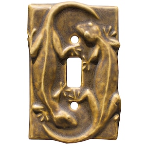 Puede incluir: Una placa de interruptor de luz de color bronce con un diseño en relieve de dos lagartos. Los lagartos están colocados alrededor de la abertura del interruptor, con detalles intrincados y una superficie texturizada. La placa tiene dos orificios para tornillos.