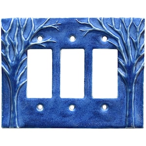 May include: A blue ceramic triple light switch cover with a tree design. The trees are white and are on the left and right sides of the cover. The cover has three rectangular openings for the switches and three small holes for screws.
