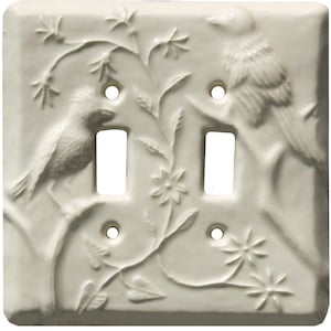 Könnte beinhalten: Doppelte Lichtschalterabdeckung aus cremefarbener Keramik mit einem erhabenen Reliefdesign von zwei Vögeln auf Ästen mit Blättern und Blumen. Sie hat zwei Schalteröffnungen und vier Schraubenlöcher.