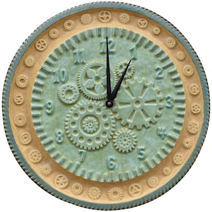 May include: Round wall clock with a steampunk design. The clock face is a muted green with raised gears and numbers. The outer ring is tan with small gear details, and the edge is a darker blue-green. The clock hands are black.