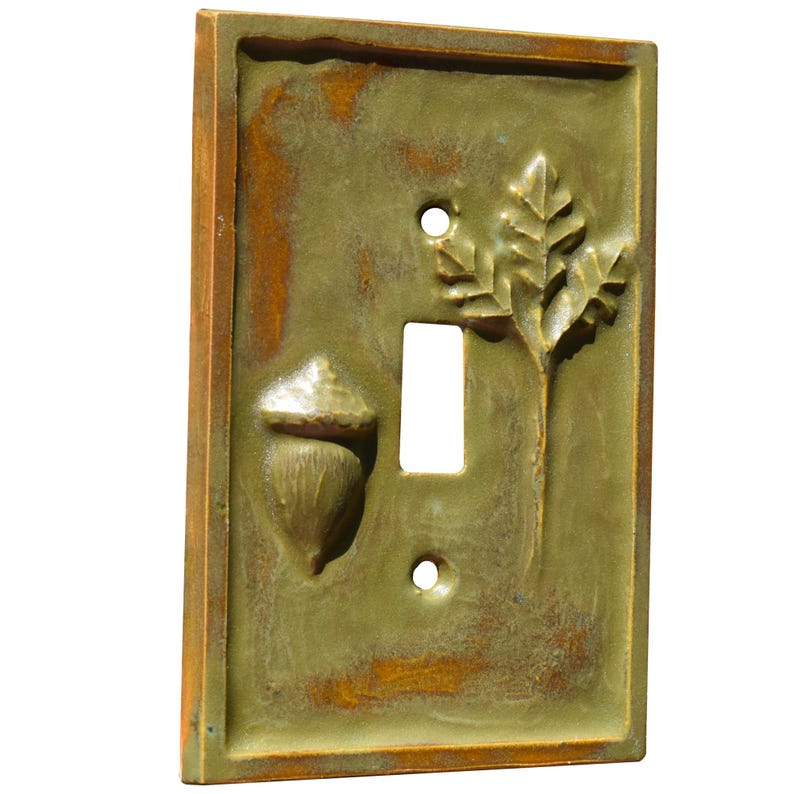 Puede incluir: Una tapa de interruptor de luz de cer&aacute;mica verde con un dise&ntilde;o de bellota y hoja de roble. La tapa del interruptor tiene un aspecto r&uacute;stico y desgastado.