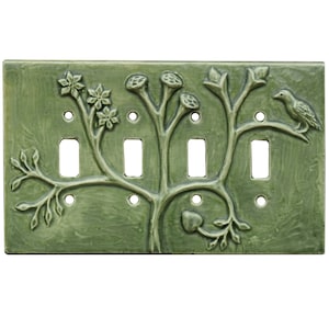 May include: A green ceramic light switch cover featuring a raised design of a stylised tree with leaves, flowers, and a bird. The switch cover has three toggle switches.