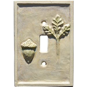 May include: A ceramic light switch cover with a nature-inspired design. The cover is a rectangular shape with a neutral color. It features an acorn and a leaf design, with a single switch opening and two screw holes.