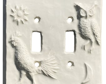 May include: A square, off-white double light switch plate featuring embossed relief designs. The plate displays a rooster on the lower left, a sun in the upper left, an owl on the upper right, and a crescent moon in the lower right corner.