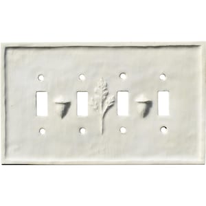 Könnte beinhalten: Eine weiße Keramik-Lichtschalterplatte mit vier Schalteröffnungen. Die Platte hat geprägte Eichel- und Blattmotive. Die Platte ist rechteckig mit abgerundeten Ecken und Befestigungslöchern.