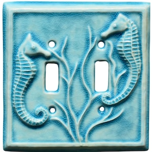 May include: A light blue ceramic double light switch cover with a textured, crackled finish. Two seahorses are sculpted on the cover, flanking the switch openings. The design includes stylized coral and plant life.