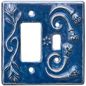 May include: A rectangular ceramic light switch plate in a deep blue glaze with a glossy finish. The plate features embossed floral motifs with white highlights, including a vertical stem on the left and curved flower buds on the right.