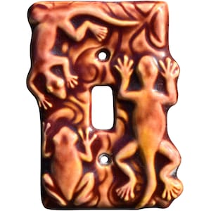 Puede incluir: Cubierta decorativa para interruptor de luz con un diseño en relieve que presenta lagartos y una rana en tonos marrones y naranjas. La cubierta tiene una abertura rectangular y dos orificios para tornillos.
