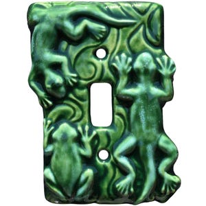 Può includere: Copri interruttore in ceramica verde con design a rilievo di rane e foglie. Il coperchio ha una finitura lucida e un'apertura rettangolare per l'interruttore. Sono visibili due fori per le viti.