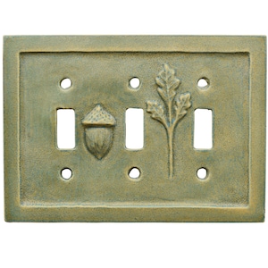 以下が含まれることがあります： くすんだ緑色の装飾的な長方形のスイッチプレート。プレートには3つのスイッチ開口部があり、ドングリとオークの葉のデザインで飾られています。プレートはテクスチャ加工された表面を持っています。