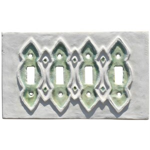 May include: A ceramic light switch plate with four toggle switch openings. The plate is white with a raised, ornate design in shades of green. The design features repeating shapes and diamond accents.