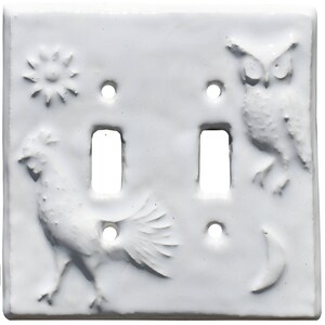 May include: A white double light switch plate featuring embossed relief designs. The surface displays a cockerel on the left, an owl on the right, a sun in the upper left corner, and a crescent moon in the lower right corner with two switch openings.