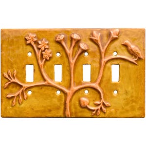 May include: A decorative light switch cover with a nature-inspired design. The cover features a raised relief of a tree with flowers, branches, and a bird. The color is a warm, golden yellow with brown accents, and it has four switch openings.