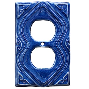 Könnte beinhalten: Eine leuchtend blaue Keramik-Doppelsteckdosenabdeckung mit einem dekorativen, fließenden Design. Sie hat zwei kreisförmige Öffnungen und ein kleines Schraubenloch. Das Design umfasst geschwungene Linien und Wirbel, mit Schnörkeldetails in den Ecken.