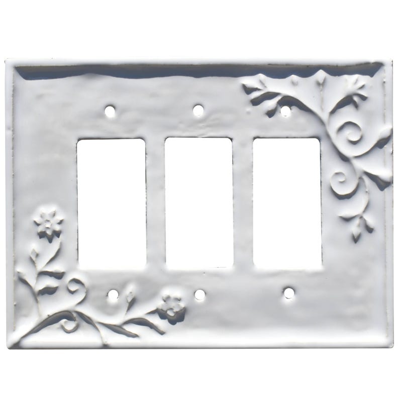 Puede incluir: Cubierta de interruptor de luz triple blanca con dise&ntilde;o floral y de vid en relieve. La cubierta tiene tres aberturas rectangulares para los interruptores. Los elementos decorativos est&aacute;n en las esquinas y los lados.