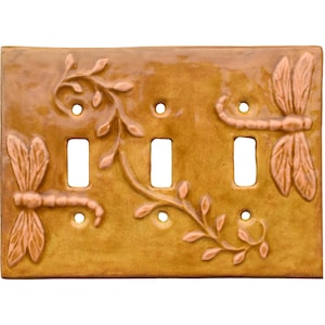May include: A rectangular ceramic light switch plate in a glossy mustard yellow finish. The plate features three vertical toggle switch cutouts and six screw holes. Embossed relief designs of two dragonflies and a leafy vine decorate the surface.