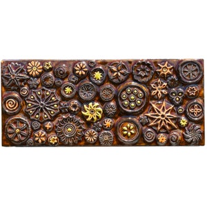 Puede incluir: Un azulejo de cerámica marrón con un mosaico de formas en relieve texturizadas en tonos marrones, dorados y amarillos. Las formas incluyen círculos, estrellas y flores.