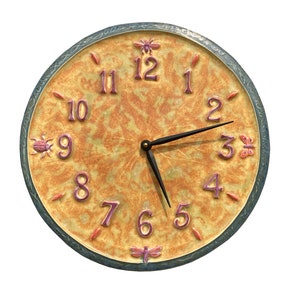 Puede incluir: Un reloj redondo de cerámica con fondo amarillo y naranja. La esfera del reloj tiene números romanos y una aguja de hora y minutos negra. El reloj está decorado con pequeños insectos coloridos.