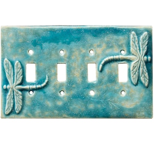 May include: A turquoise ceramic light switch plate with four switch openings. The plate features raised dragonfly designs on each end, with a textured, speckled finish. The dragonfly wings are detailed with delicate lines.