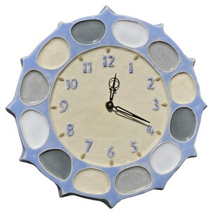 May include: A ceramic wall clock with a light blue scalloped edge and a cream-colored face. The clock features blue numerals and black hands. The outer edge has a series of oval-shaped, multi-colored stones. A decorative piece for any room.