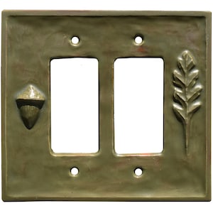 Peut inclure: Une plaque d'interrupteur double en finition vert olive sourd avec une surface texturée. La plaque présente un relief de gland sur le côté gauche et un relief de feuille de chêne sur le côté droit, avec quatre trous de vis pour la fixation.