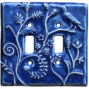 May include: A square, deep blue ceramic double light switch plate featuring a raised, embossed relief design of a bird perched on a branch surrounded by intricate swirling vines, leaves, and small flowers across the entire surface.