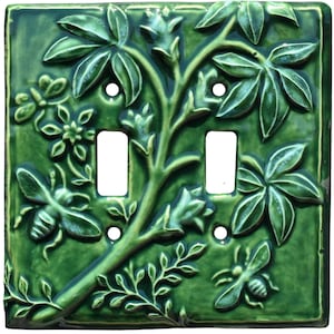 May include: Green ceramic double light switch cover with a raised floral and bee design. The cover features two toggle switch openings and four screw holes. The design includes leaves, flowers, and two bees.