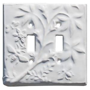 May include: A white double light switch plate featuring a raised, embossed botanical design with leaves, flowers, and two bees. The rectangular cover includes two vertical rectangular cutouts for switches and four small circular screw holes.