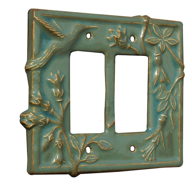 May include: A teal ceramic double light switch cover with a hummingbird and floral relief design. The cover has a glossy finish and gold accents. The hummingbird is perched on a branch, and the floral elements include flowers and leaves.