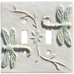May include: A square double light switch wall plate in an off-white finish featuring a raised relief design of two dragonflies and a central flower on a vine. The plate includes two rectangular cutouts for switches and four screw holes.