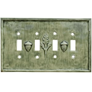 May include: A green ceramic light switch plate with a textured surface. The plate features three toggle switch openings and two acorn and one leaf design. The plate has a rectangular shape.