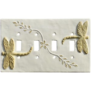 May include: A cream-coloured light switch plate featuring two dragonflies and leafy vines in a light tan hue. The plate has four switch openings and small circular screw holes, with a decorative design.