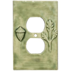 May include: A green ceramic outlet cover with an acorn and leaf design. The cover has two circular openings for outlets and a small hole for a screw. The design is embossed and adds a natural, rustic touch to any room.