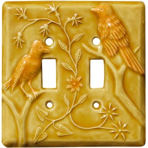 May include: A square, mustard-yellow ceramic light switch plate featuring a raised relief design of two birds perched on branches. A central vine with leaves and a small flower separates the two rectangular switch openings and four screw holes.