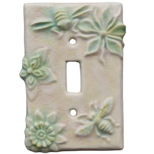 Puede incluir: Una placa de interruptor de luz rectangular de cerámica con un diseño en relieve de dos abejas y dos flores. La superficie tiene un color base blanco roto mate con suaves acentos difuminados de color verde pálido y amarillo.