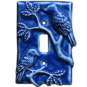 Op de afbeelding: Een blauwe keramische lichtschakelaar met twee vogels die op takken zitten. De vogels zijn in een lichtblauwe glazuur en de takken in een donkerdere blauwe glazuur.
