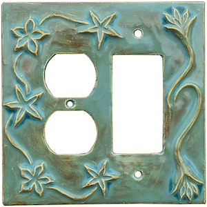 以下が含まれることがあります： 正方形のターコイズ色のセラミック製ライトスイッチカバー。コンセントが2つとスイッチ用の穴があります。カバーは、対照的なゴールドカラーの隆起した花柄と星型のデザインで装飾されています。全体的なデザインは手作り感があります。
