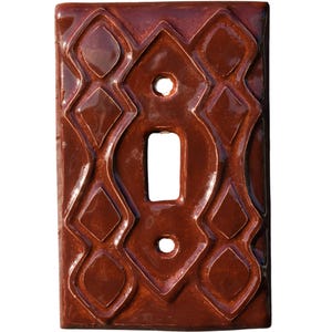 May include: A rectangular, brown ceramic light switch cover with a glossy finish. The cover features a central opening for the switch and decorative diamond and curved line patterns. Two small holes are above and below the switch opening.