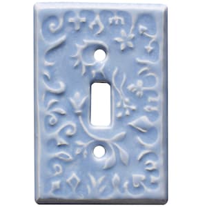 Könnte beinhalten: Eine hellblaue Keramik-Lichtschalterabdeckung mit erhabenem Blumen- und Scroll-Design. Die Abdeckung hat eine einzelne Kippschalteröffnung und zwei Schraubenlöcher. Das dekorative Design ist in einem helleren Blauton gehalten.